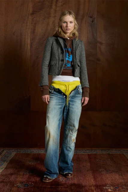 Woman in fashionable pose wearing a gray tweed jacket, brown hoodie, and ripped jeans stands against a wood backdrop and patterned rug.