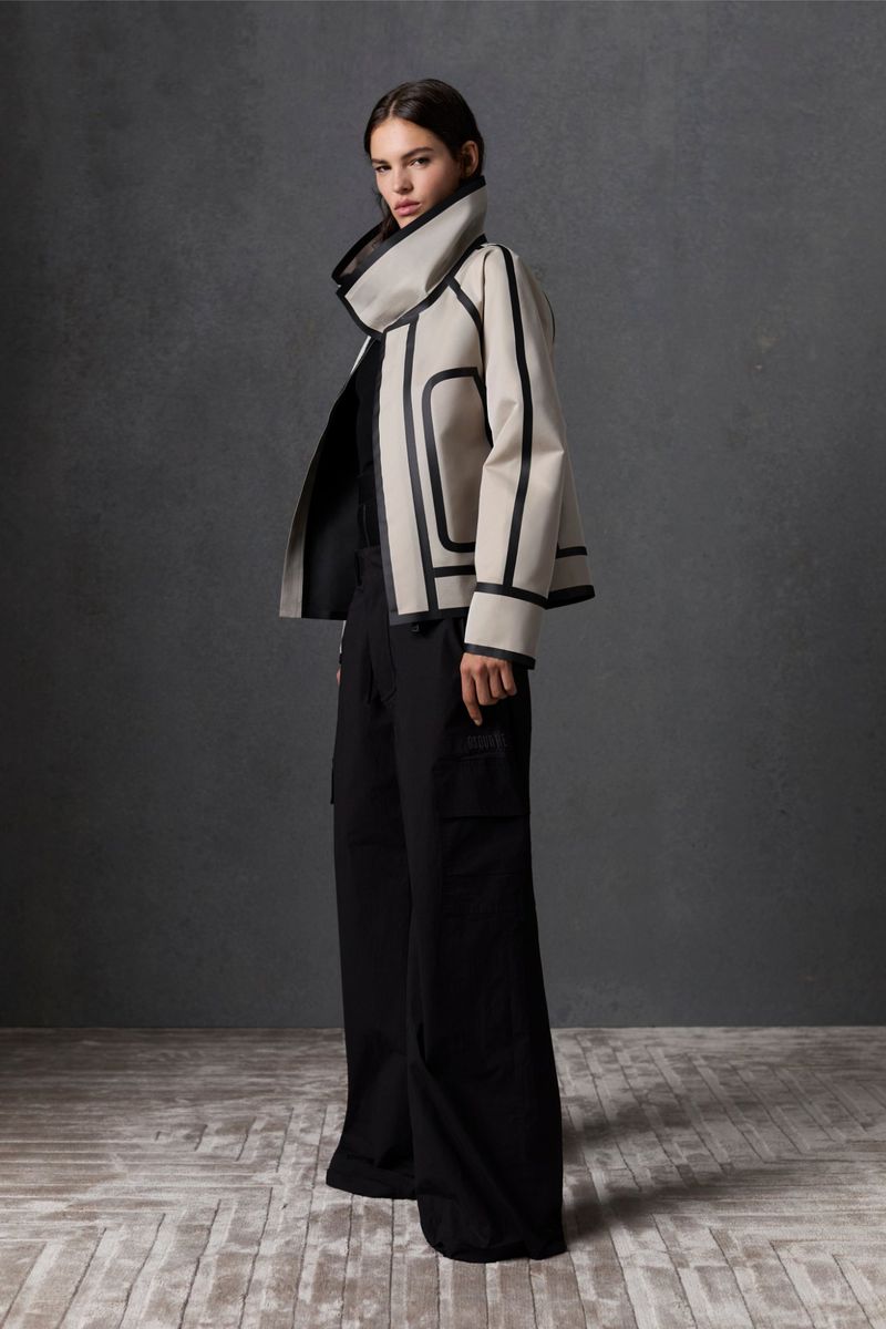 Fashionable adult female model wearing a cream-colored jacket with black trim and black pants posing in front of a dark grey background.