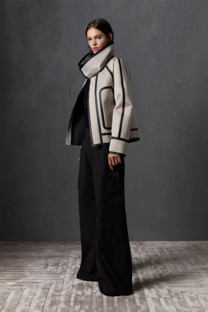 Fashionable adult female model wearing a cream-colored jacket with black trim and black pants posing in front of a dark grey background.