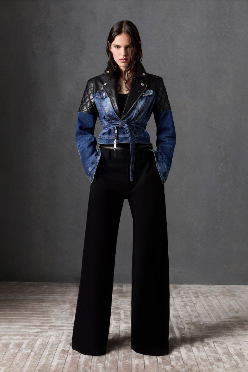 Full-length shot of an adult woman in a denim jacket with leather shoulders and black wide-leg trousers, standing against a grey backdrop.