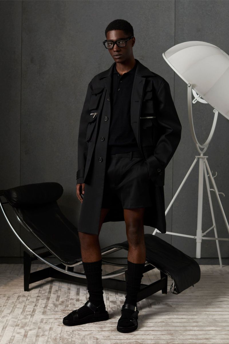 A Black male model poses in an all-black casual outfit of coat, shorts, polo shirt, socks, and sandals in a minimalist studio.