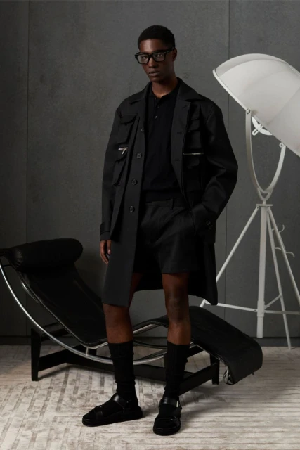 A Black male model poses in an all-black casual outfit of coat, shorts, polo shirt, socks, and sandals in a minimalist studio.