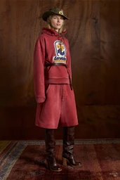 Model in red hoodie and matching shorts wearing tall brown boots and a hat against a wooden wall background.