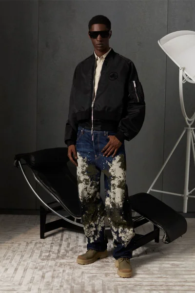 Model wearing black bomber jacket, patterned jeans, and sunglasses standing in a modern studio setting with a lounge chair