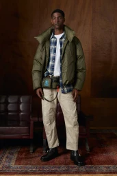Model in olive puffer jacket, plaid shirt, and cream trousers standing in an interior space