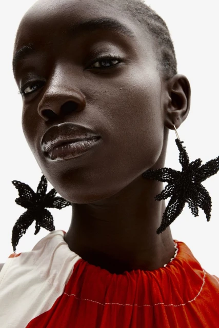 Close-up of a Black woman with glossy lips wearing large, black, star-shaped earrings and a red and white top against a white background.
