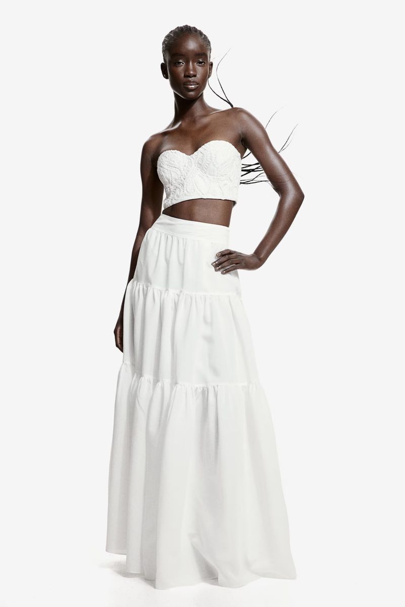 Elegant Black woman poses in a white strapless lace crop top and flowing tiered white skirt against a bright white background.