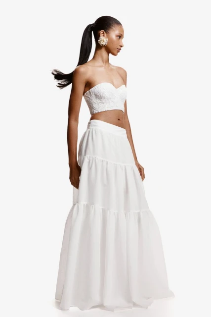 Studio shot of a Black female model in a white two-piece dress, featuring a cropped top and a tiered maxi skirt, against a white background.