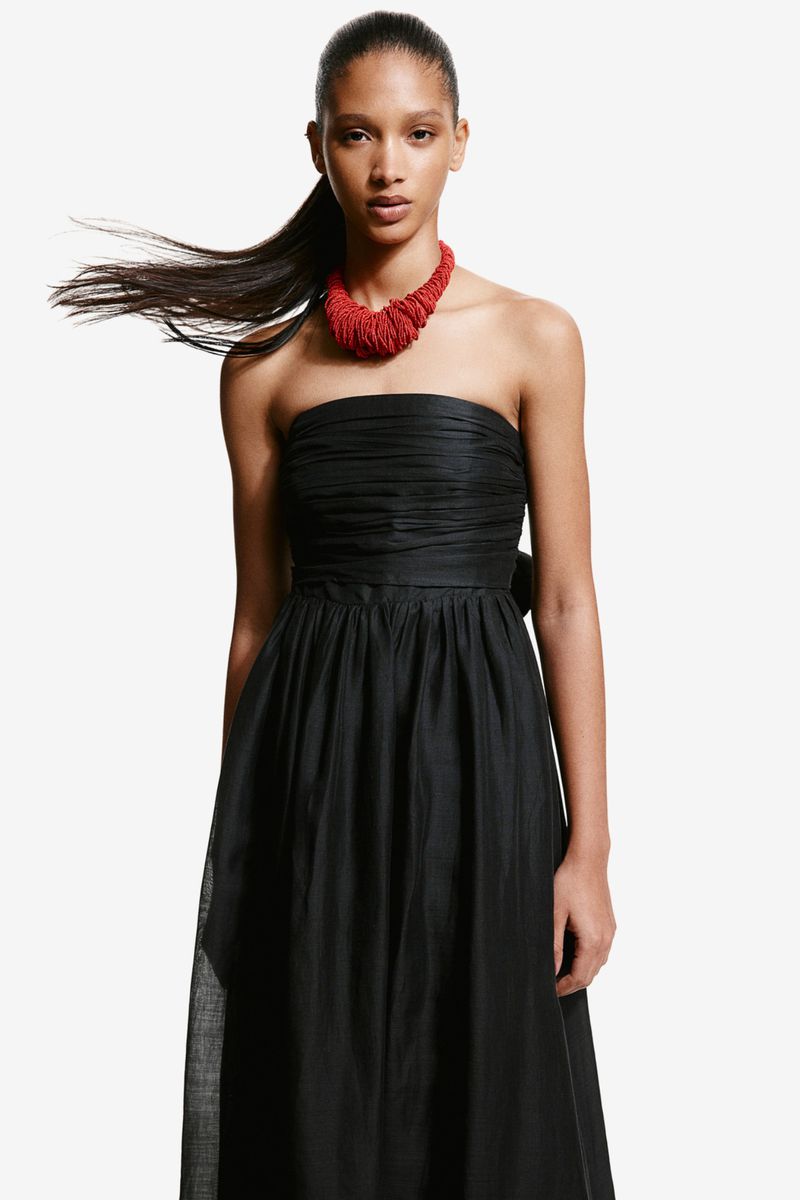 A woman poses in a black strapless dress with a red necklace against a white background, showcasing fashion and elegance.