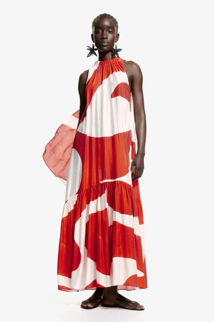Elegant Black woman wearing red-and-white halter dress and earrings poses in front of a white backdrop in a full studio shot.