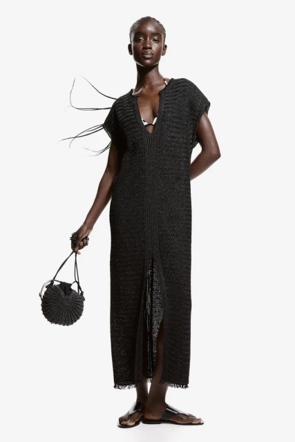 An elegant, dark-skinned woman models a long black knit dress with a circular woven bag against a bright white studio backdrop.