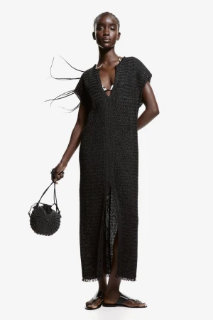 An elegant, dark-skinned woman models a long black knit dress with a circular woven bag against a bright white studio backdrop.