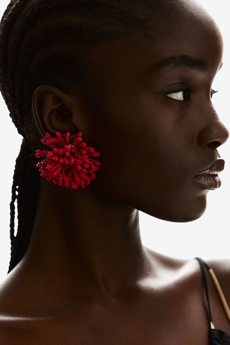 Close-up of a dark-skinned woman wearing a large, bright red pompom earring, looking to the side against a bright white background.