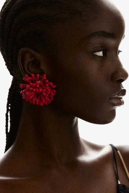 Close-up of a dark-skinned woman wearing a large, bright red pompom earring, looking to the side against a bright white background.