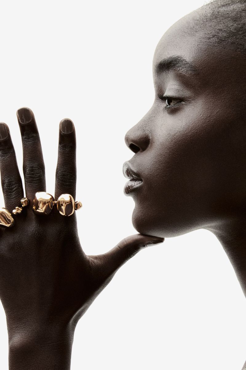 Close-up profile of a Black woman wearing multiple gold rings, set against a bright white backdrop in a studio setting.