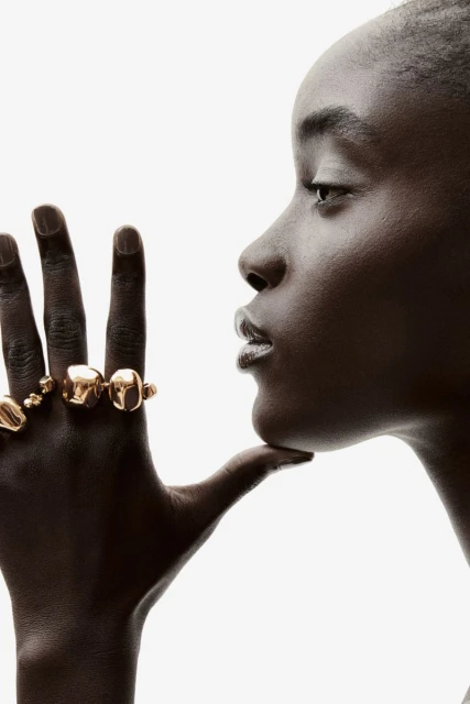 Close-up profile of a Black woman wearing multiple gold rings, set against a bright white backdrop in a studio setting.