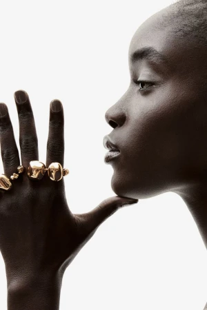 Close-up profile of a Black woman wearing multiple gold rings, set against a bright white backdrop in a studio setting.