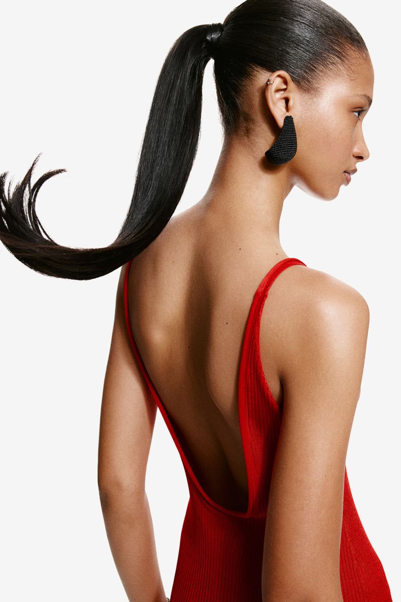 Portrait of an adult woman with black hair in a ponytail and a red backless top against a white background in a studio setting.