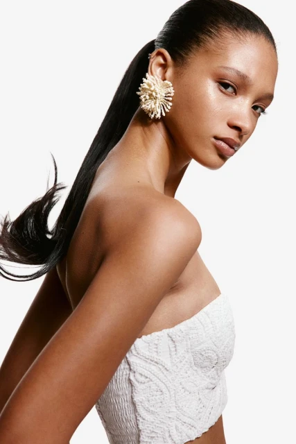 An adult woman with brown skin and black hair in a white textured top poses against a white background wearing a large floral earring.