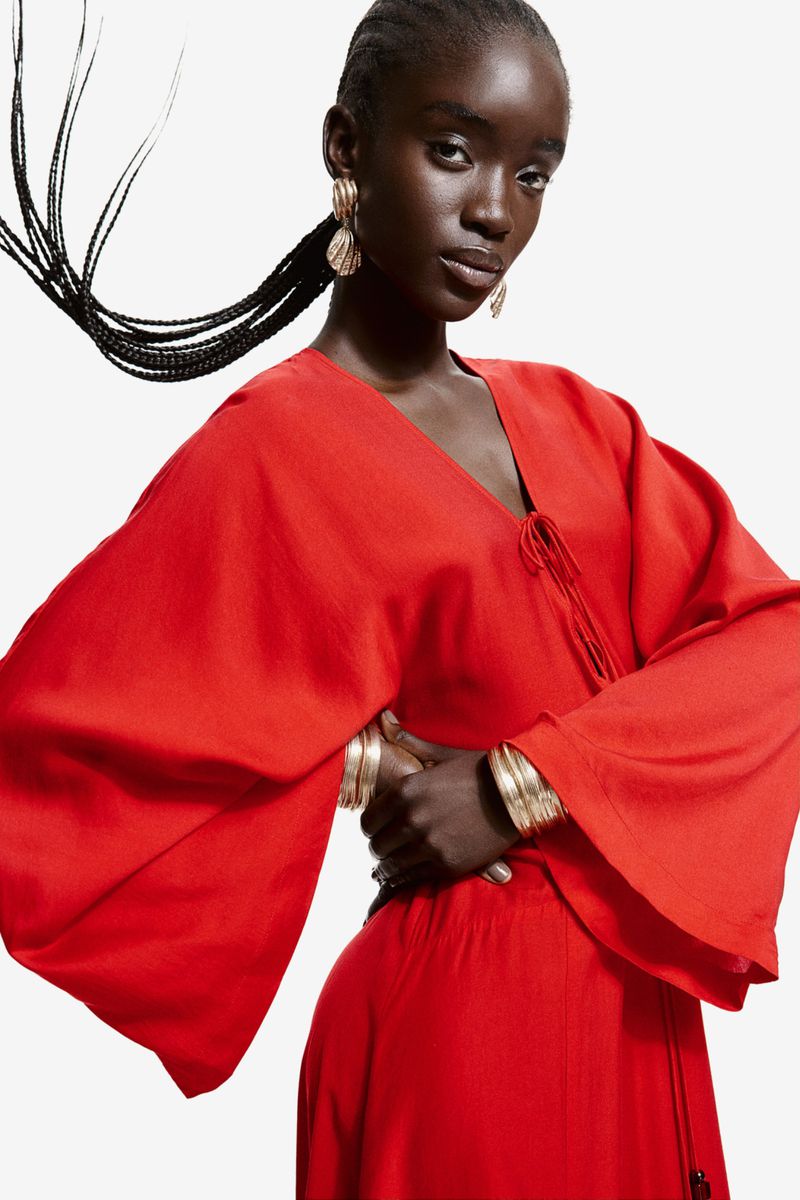 Model in a red dress with wide sleeves wearing gold earrings and stacked bracelets against a white background