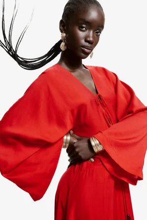 Model in a red dress with wide sleeves wearing gold earrings and stacked bracelets against a white background