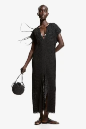 Model in a long black textured dress holding a dark woven handbag against a plain white background.