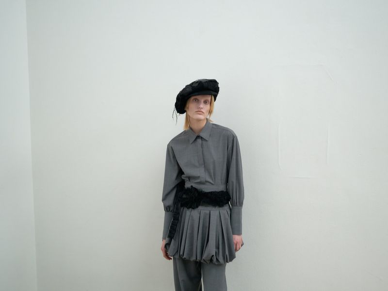 Blonde model poses in gray shirt and pleated skirt with black belt against a white wall in a fashion editorial shot.