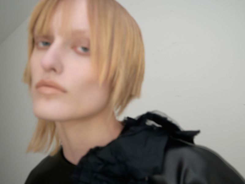 Close-up of an adult female with short blonde hair and a black frilled collar, standing in an indoor environment with a neutral background.
