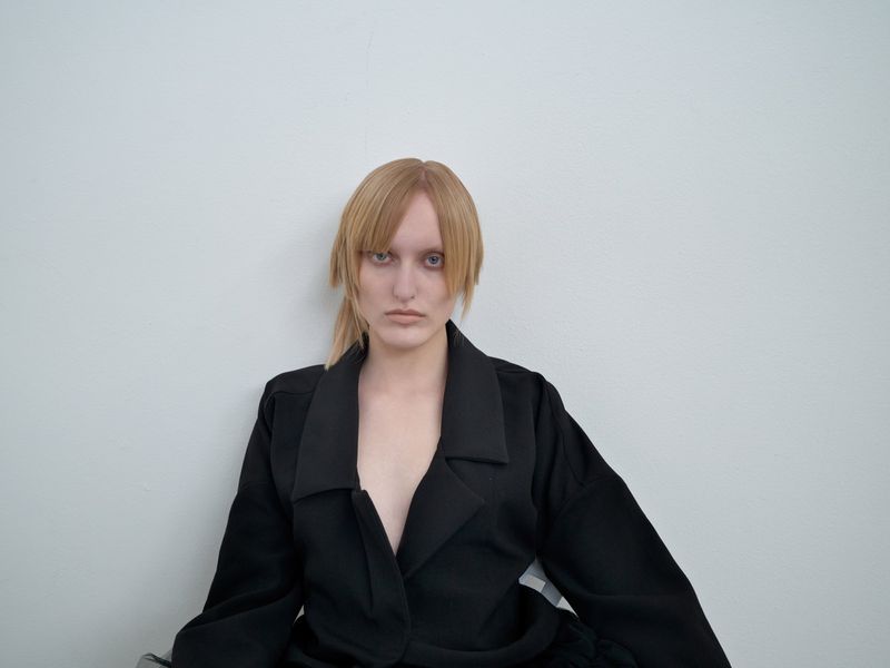 Portrait of a blonde woman with bangs wearing a black jacket in front of a white wall, showcasing fashion and simple background.