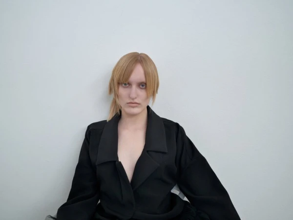 Portrait of a blonde woman with bangs wearing a black jacket in front of a white wall, showcasing fashion and simple background.