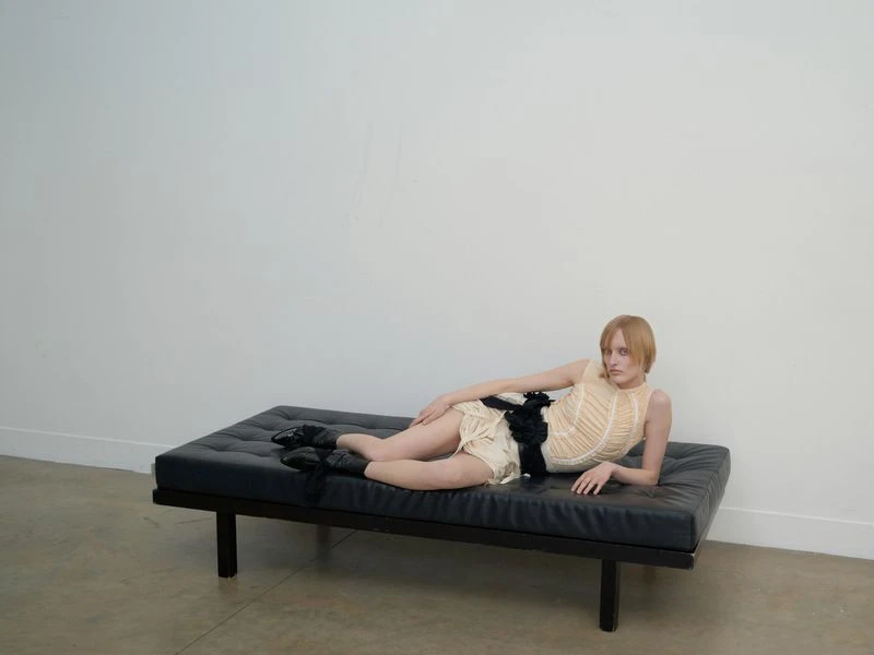 An adult female model with short blond hair poses on a black modern daybed in a minimalist indoor space with white walls.