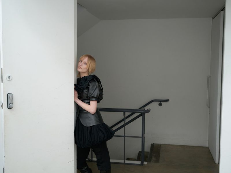 Fashion photo of a blonde woman in a black outfit, leaning against a white door in an indoor stairwell, captured in a studio setting.