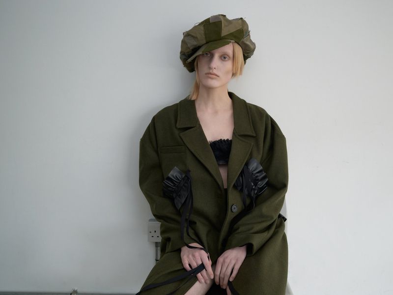 Blonde-haired woman in an olive coat and cap poses against a white wall, showcasing modern fashion with a hint of vintage flair.