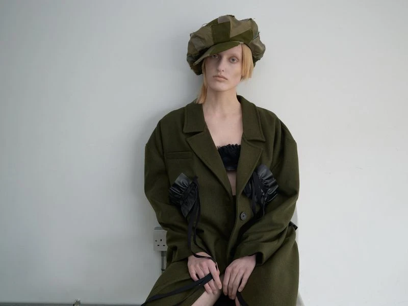 Blonde-haired woman in an olive coat and cap poses against a white wall, showcasing modern fashion with a hint of vintage flair.