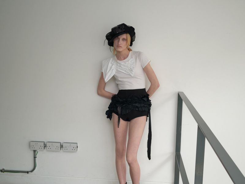 A model poses in a studio, wearing a white t-shirt, black frilly bottoms, and a wide-brimmed black hat. The background is a plain white wall.