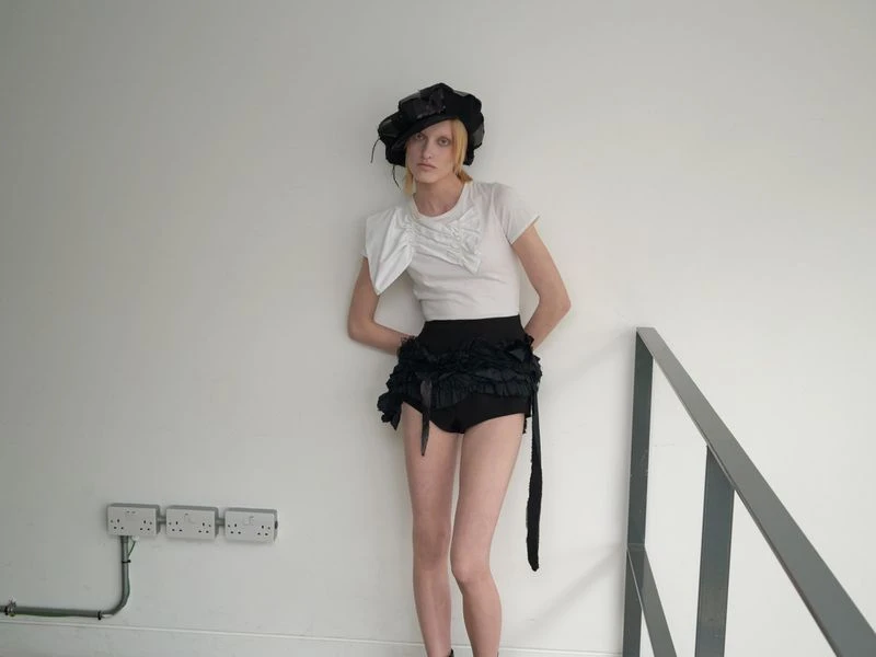 A model poses in a studio, wearing a white t-shirt, black frilly bottoms, and a wide-brimmed black hat. The background is a plain white wall.