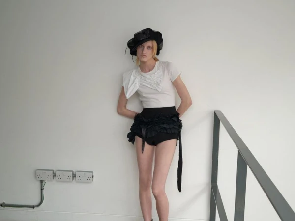 A model poses in a studio, wearing a white t-shirt, black frilly bottoms, and a wide-brimmed black hat. The background is a plain white wall.