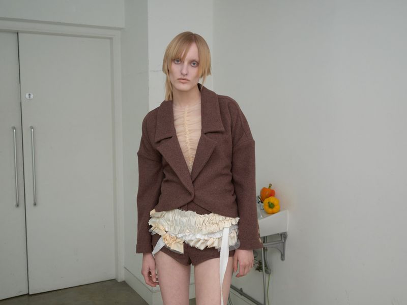 A fashion shot presents a model in a brown jacket, shorts, and ruffled embellishments, posed indoors against a backdrop of a white wall and door.
