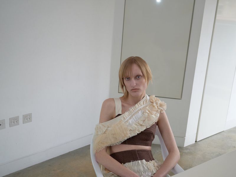 Fashion shot of a woman in a brown and beige outfit with ruffles, sitting in a bright white room with simple decor and a neutral expression.