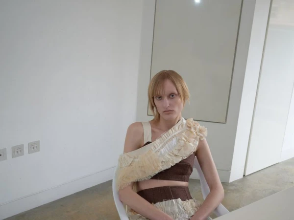 Fashion shot of a woman in a brown and beige outfit with ruffles, sitting in a bright white room with simple decor and a neutral expression.