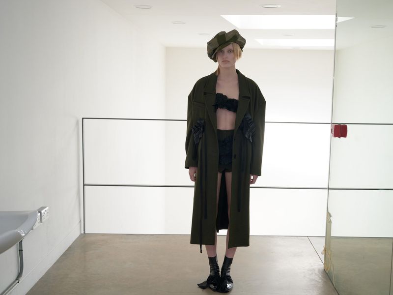 A woman wearing an olive-green coat and matching cap stands in a bright, minimalist indoor setting with a sink and mirrors.
