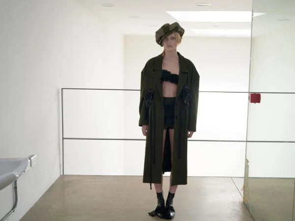 A woman wearing an olive-green coat and matching cap stands in a bright, minimalist indoor setting with a sink and mirrors.