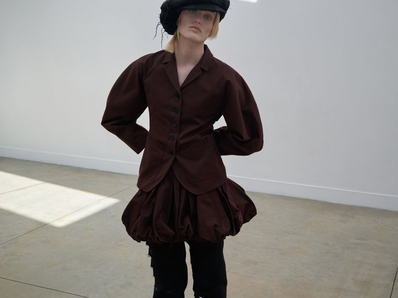 Woman in a brown jacket and puffy skirt poses in front of a white wall with light concrete flooring in a fashion shot.