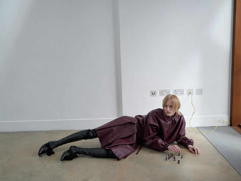 A person in a maroon outfit lies on the floor with dark boots and plays with chess pieces in front of a white wall with electrical outlets.