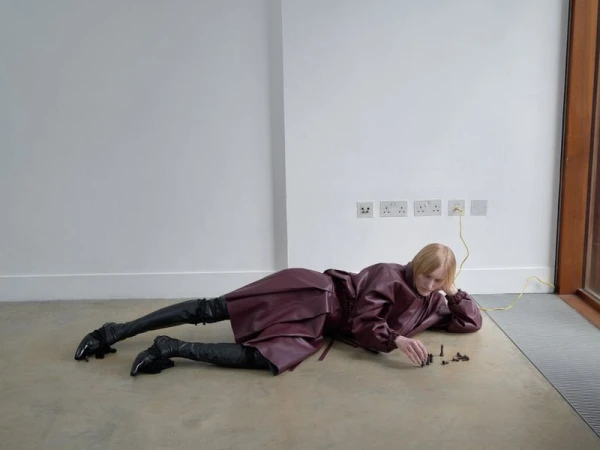 Woman in burgundy coat and black boots lies on the floor arranging chess pieces near an electrical outlet in a minimalist room.