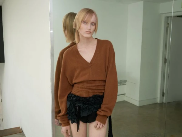 Fashion portrait of a slender model with fair skin wearing a brown sweater and black ruffled shorts in a minimalist photo studio.