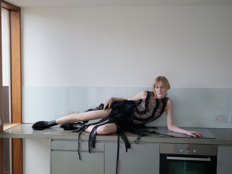 A slender woman with fair skin and light hair poses in a black sheer dress and boots on a kitchen counter, against a white wall.