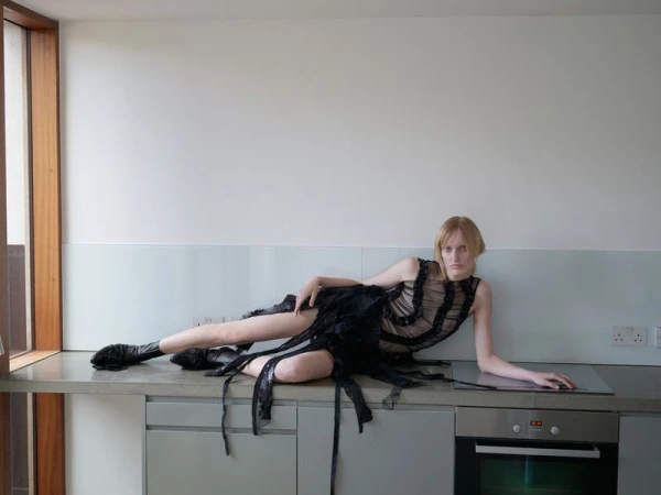 A slender woman with fair skin and light hair poses in a black sheer dress and boots on a kitchen counter, against a white wall.
