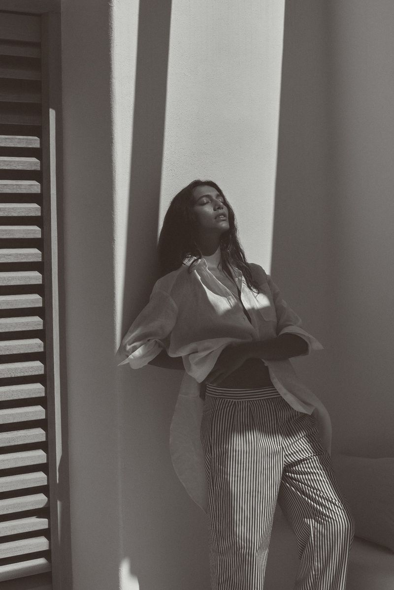 Woman in sunlight wearing an unbuttoned white blouse and striped pants leans against a white wall with a slatted door.