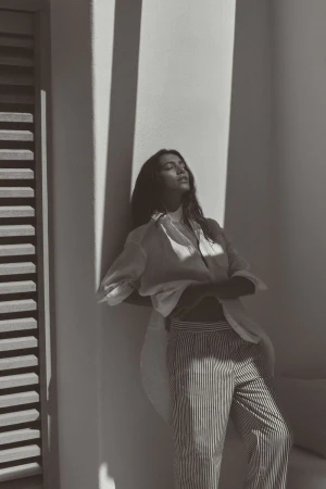 Woman in sunlight wearing an unbuttoned white blouse and striped pants leans against a white wall with a slatted door.
